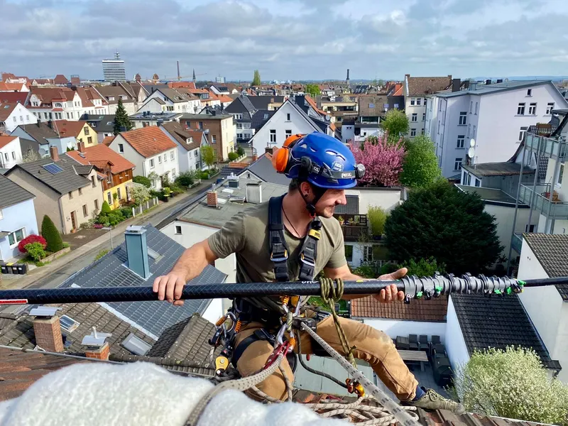 Industriekletterer mit Auffanggurt und Seilsicherung auf einem Dach über der Stadt